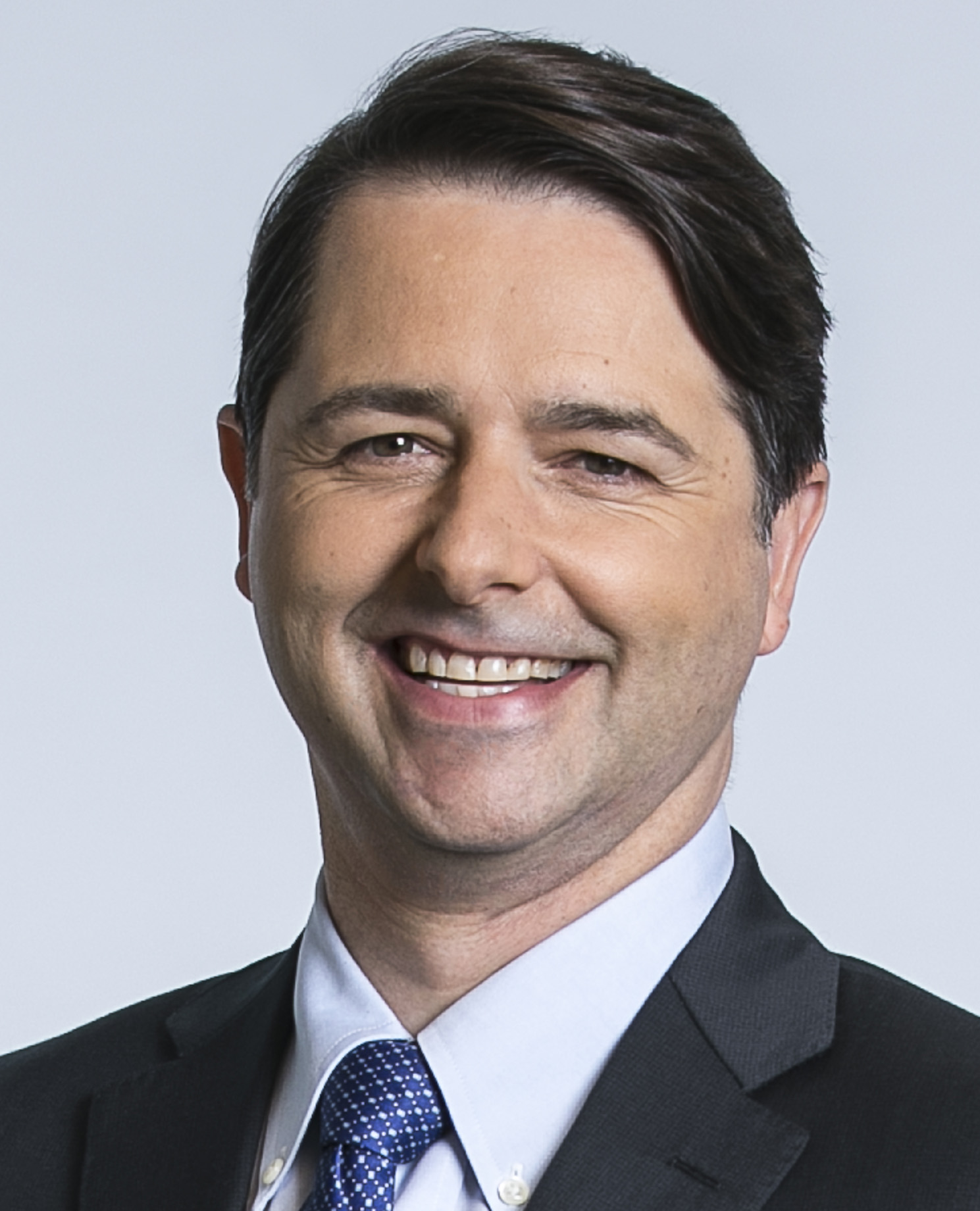 A man in a dark suit with a blue tie is smiling and looking at the camera.