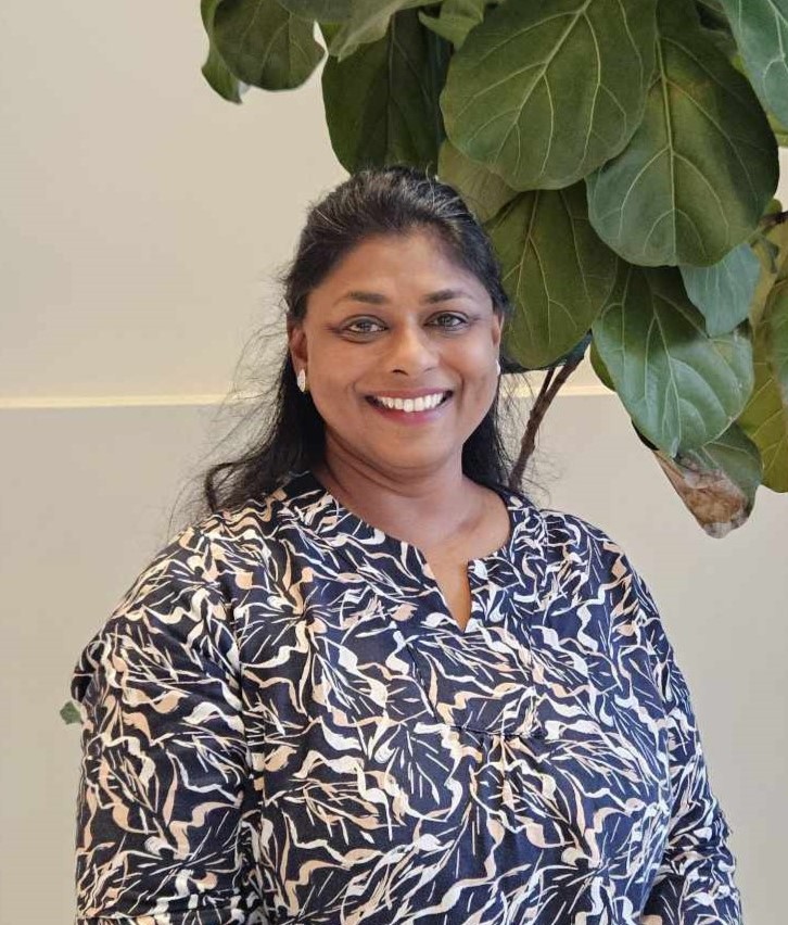 A women with a white patterned shirt is standing in front of a green bush and smiling at the camera.