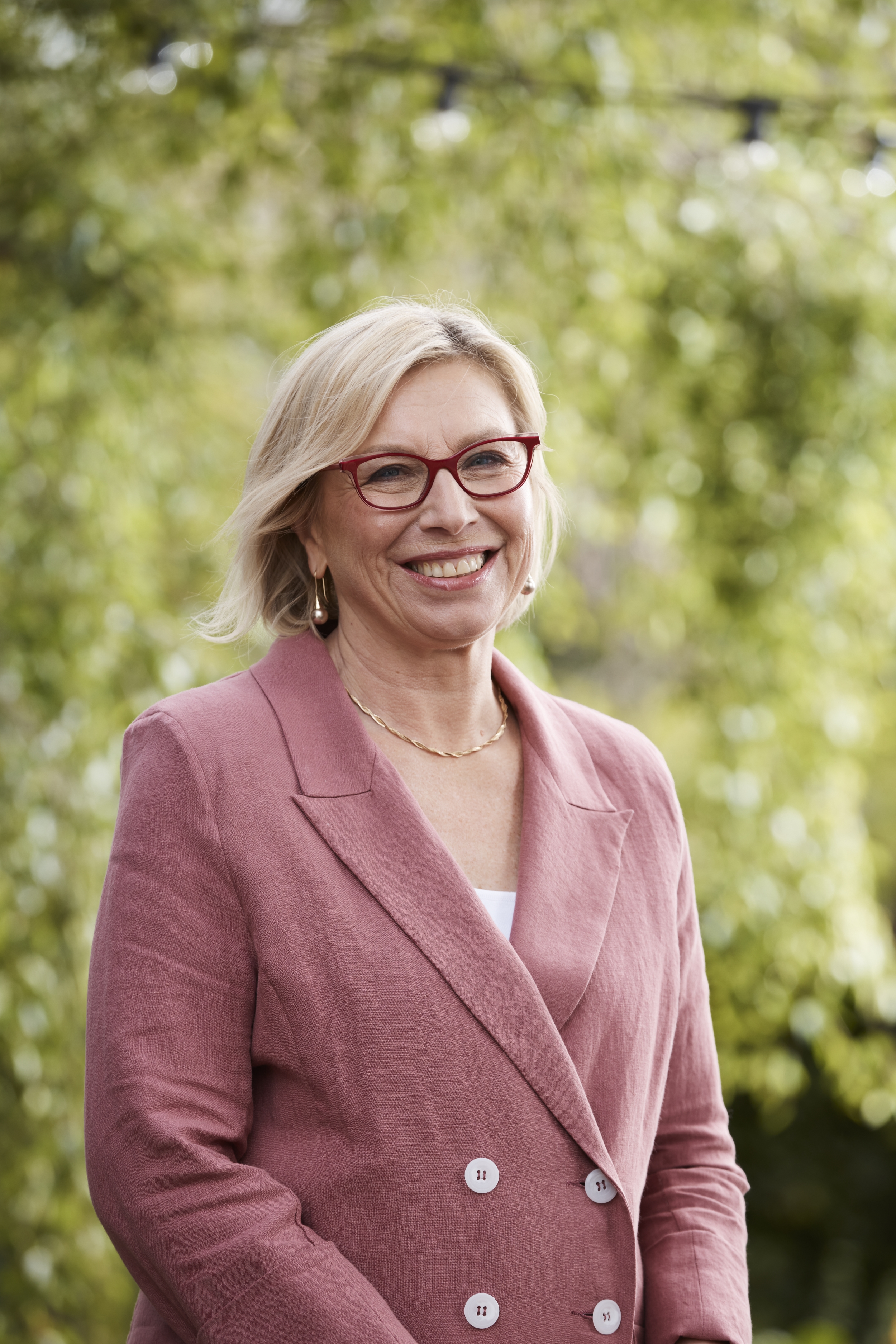 A lady with blonde hair and glasses, and wearing a light pink jacket, is standing outdoors and smiling at the camera.