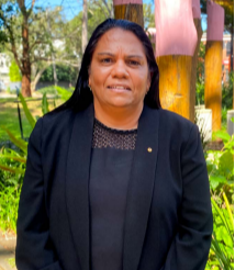 A woman is wearing a black jacket and is standing facing the camera, with her hands folded in front of her.
